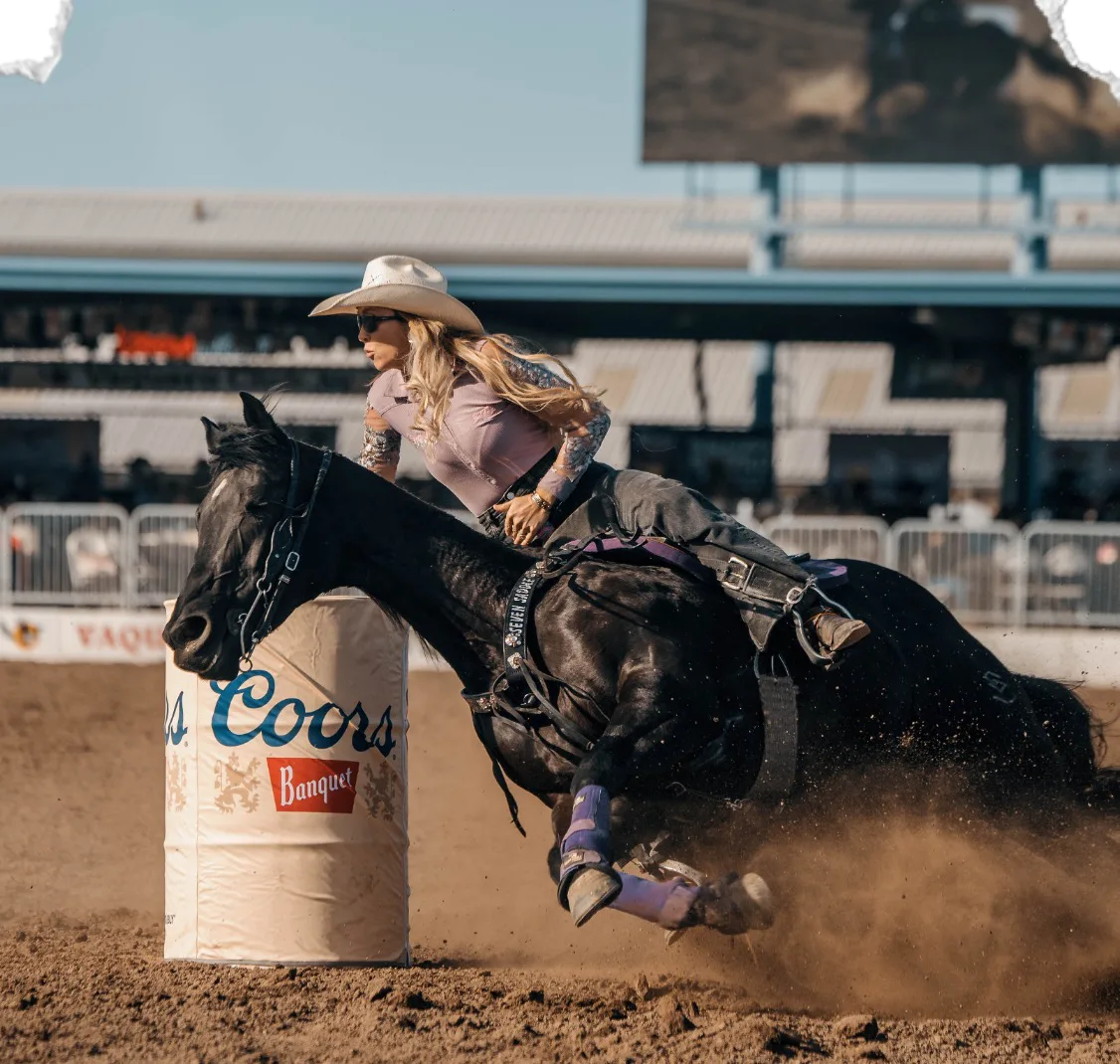 A barrel racer turns her black horse around a Coors barrel.