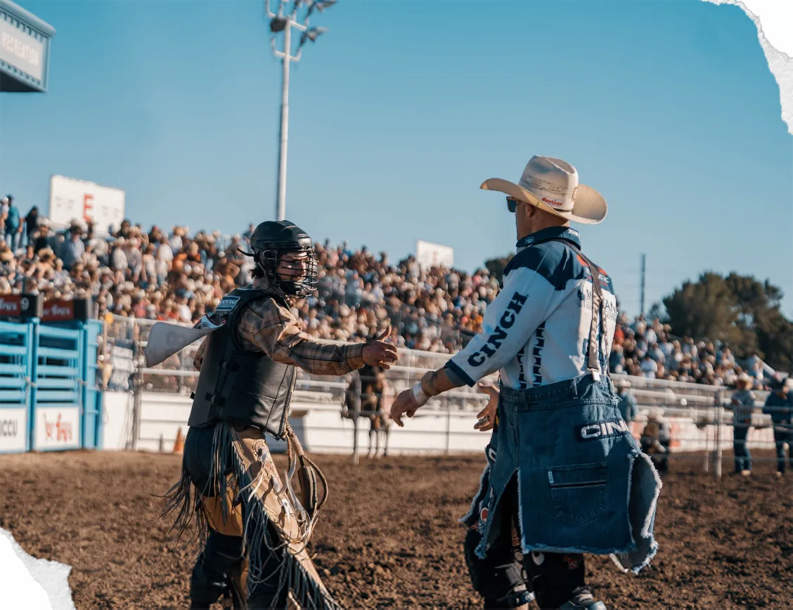 A bullfighter in a cowboy hat shakes hands with a bull rider in the arena.