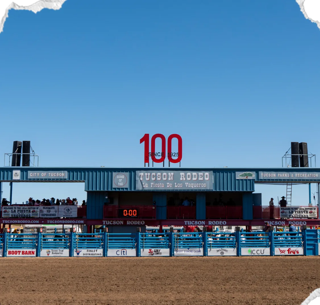The Tucson Rodeo grandstand and bucking chutes across the arena, under a red 100 sign and a clear blue sky.