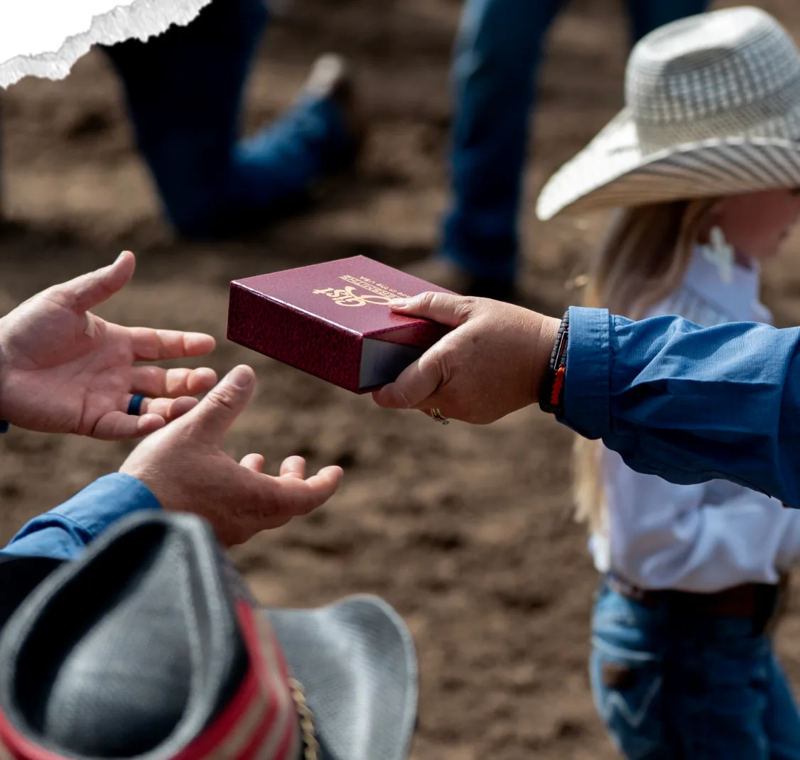 In the arena, a small maroon presentation box is handed to outstretched hands while a girl in a straw cowboy hat stands behind.