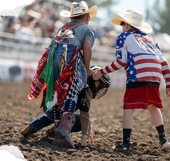 Two young Junior Rodeo competitors in the arena, one helping the other up from the dirt